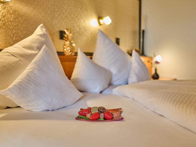 Nahaufnahme eines Hotelbetts mit weißen Kissen und dekorativem Teller mit Früchten und Pralinen.