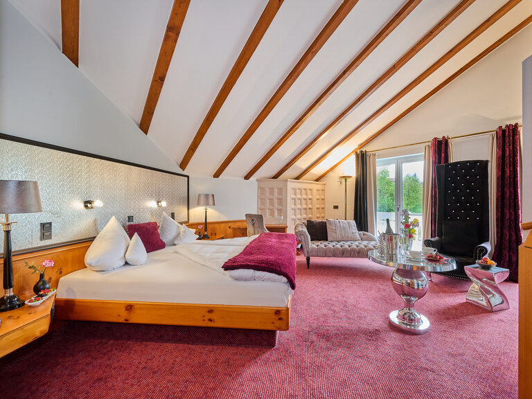 Geräumige Suite mit Dachschrägen, rotem Teppich, Doppelbett, Sitzmöbeln und Balkontür.
