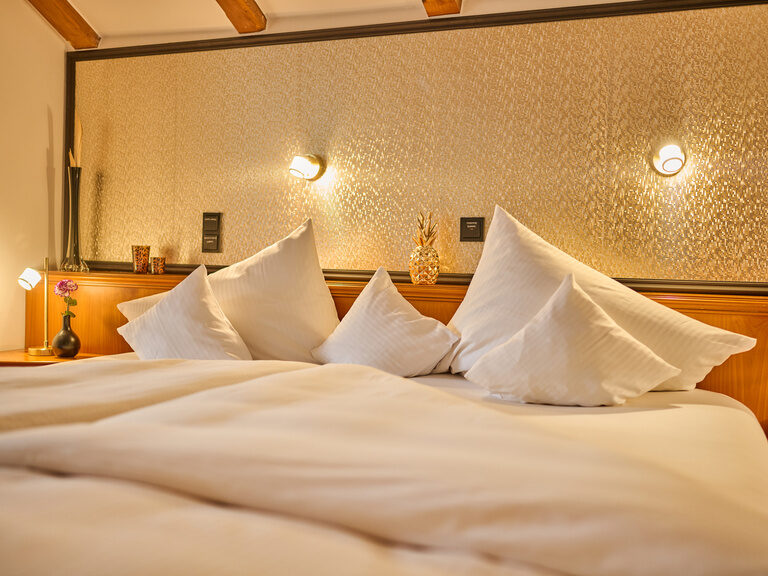 Doppelbett mit weißen Kissen, warmer Beleuchtung und goldfarbener Rückwand.