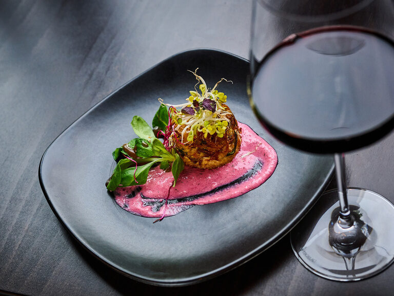 Vorspeise auf schwarzem Teller mit rosa Sauce und Rotwein im Vordergrund.