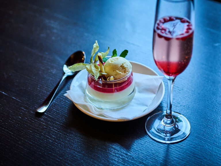 Dessert mit Eis und Früchten neben Cocktail auf dunklem Tisch.