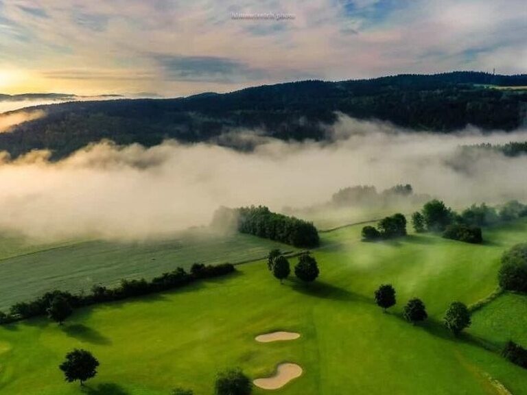 Landschaft mit Golfplatz im Morgennebel, darüber bewaldete Hügel bei Sonnenaufgang.