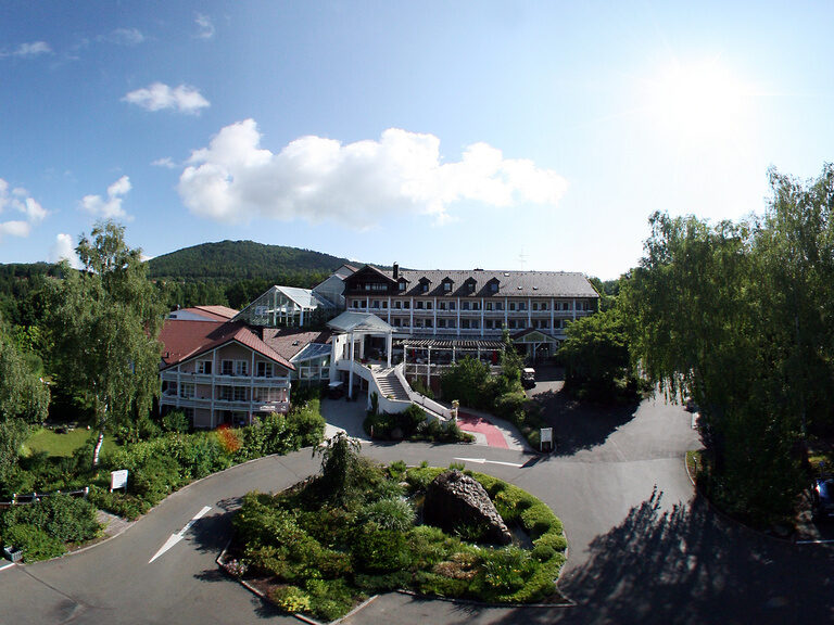 Außenansicht einer Hotelanlage mit Grünflächen, Auffahrt und bewaldeten Hügeln im Hintergrund.