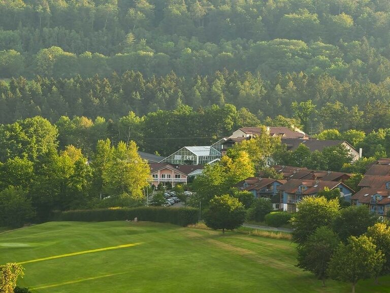 Hotelanlage mit Glasdach inmitten von Häusern am Rand eines Waldstücks.