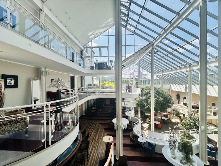 Helles Atrium mit Glasdach, weißen Balustraden, Pflanzen und Sitzgruppen auf mehreren Ebenen.