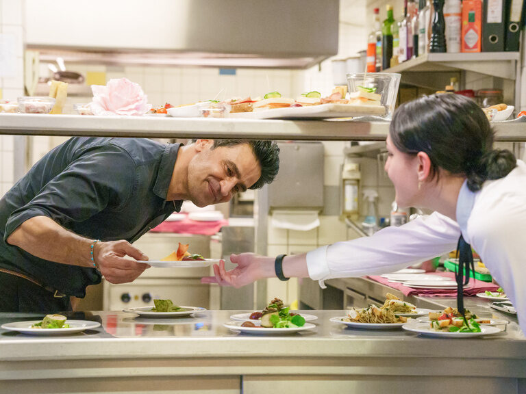 Kellnerin nimmt Teller vom Küchenpass mit lächelndem Koch entgegen.