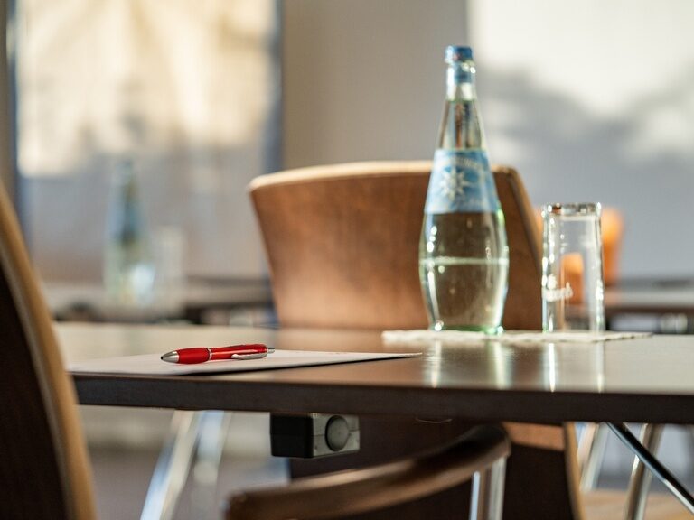 Detailaufnahme von Stuhl und Tisch mit Wasserflasche, Glas und Schreibset im Tageslicht.