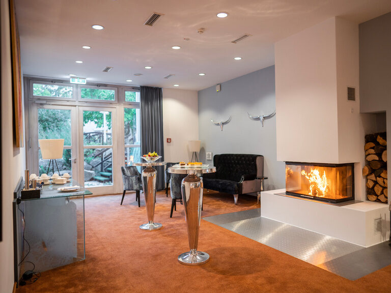 Moderner Aufenthaltsraum mit Kamin, hohen Tischen und direktem Zugang ins Freie.