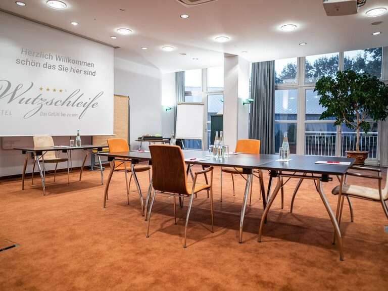 Konferenzraum mit Projektionsfläche, Tischen, Wasserflaschen und natürlichem Lichteinfall.