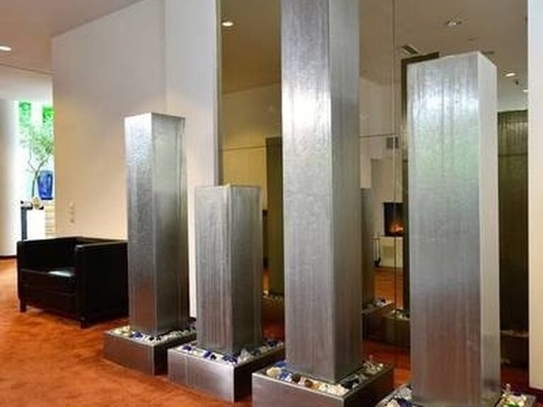 Vier hohe Wasserinstallationen aus Metall vor Spiegelwand auf rotem Teppichboden.