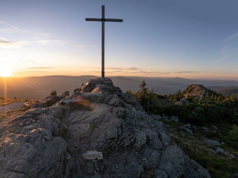 Sonnenaufgang am Gipfelkreuz mit Panoramablick auf bewaldete Berge.