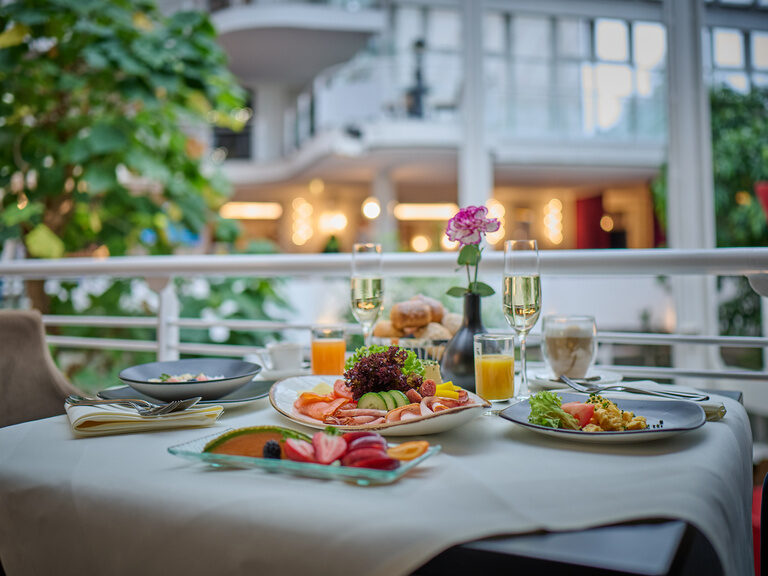 Frühstückstisch mit Aufschnitt, Eierspeisen, Saft, Kaffee und Blume im hellen Innenraum.