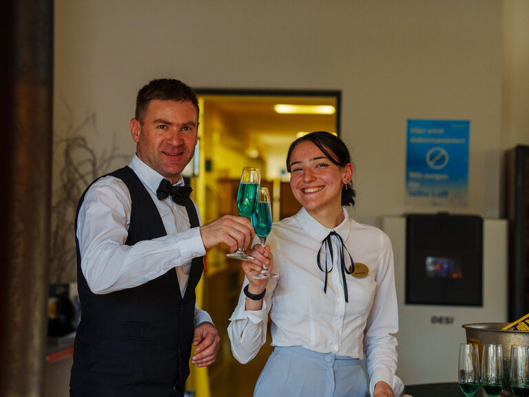 Zwei Servicekräfte stoßen lächelnd mit türkisfarbenen Sektgläsern im Eingangsbereich an.