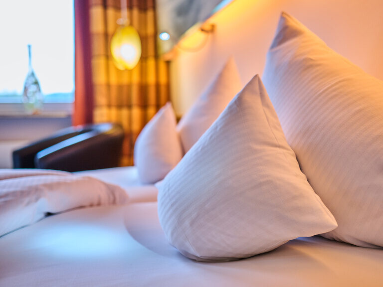 Nahaufnahme von weißen Kopfkissen auf Hotelbett, stimmungsvolle Beleuchtung im Hintergrund.