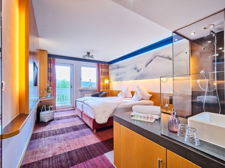 Hotelzimmer mit Doppelbett, Teppichboden, Glasdusche und Blick ins Grüne.
