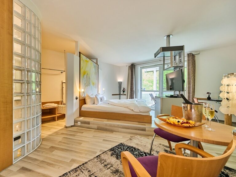 Geräumige Hotelsuite mit offenem Bad, Glastrennwand, Sitzecke und Balkonblick ins Grüne.