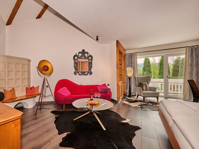 Gemütlicher Sitzbereich im Hotelzimmer mit rotem Sofa, Kuhfell-Teppich und Balkon.