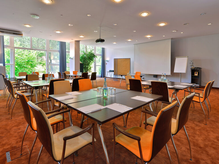 Tageslichtraum mit viereckigen Tischgruppen, Block und Stift an jedem Platz.