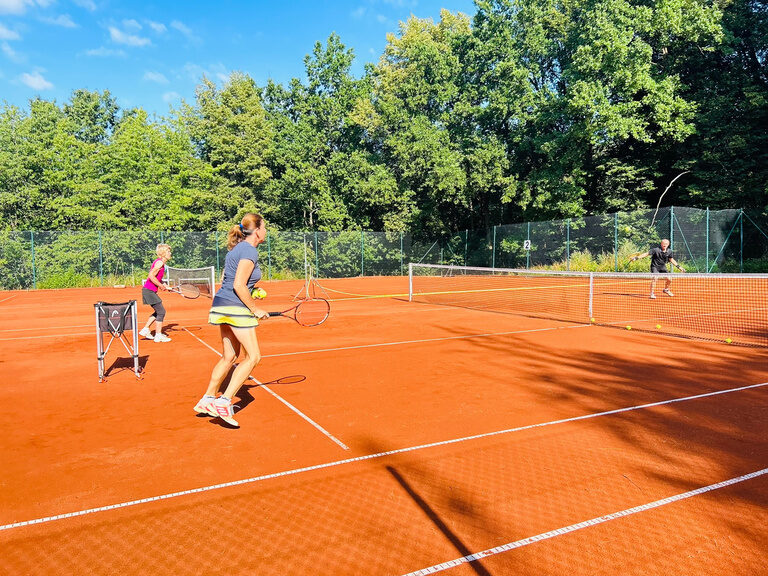 Drei Personen beim Tennistraining auf Sandplatz im Freien bei Sonnenschein.