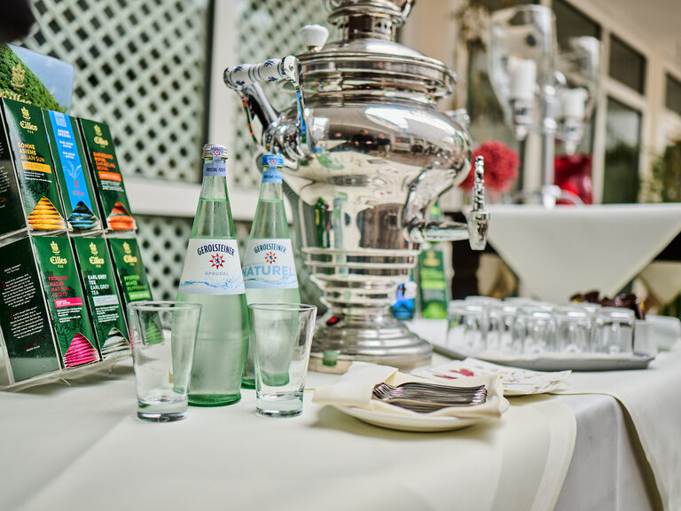 Teestation mit Gerolsteiner-Wasserflaschen und Samowar auf weiß gedecktem Tisch.
